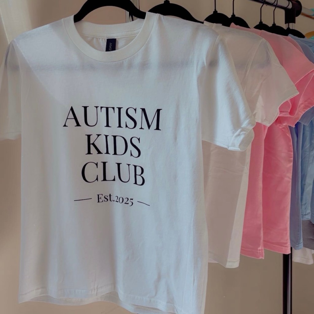 A white t-shirt featuring the text 'AUTISM KIDS CLUB' and 'Est. 2025', hanging alongside several other colored shirts.